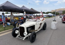 Front three-quarter view of an off-white 1932 Ford roadster hot rod with "Banger Power" hood lettering carrying passengers down the fairgrounds thoroughfare past spectator canopies.