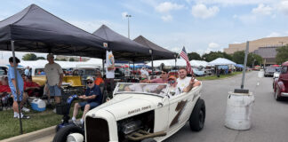 Front three-quarter view of an off-white 1932 Ford roadster hot rod with "Banger Power" hood lettering carrying passengers down the fairgrounds thoroughfare past spectator canopies.