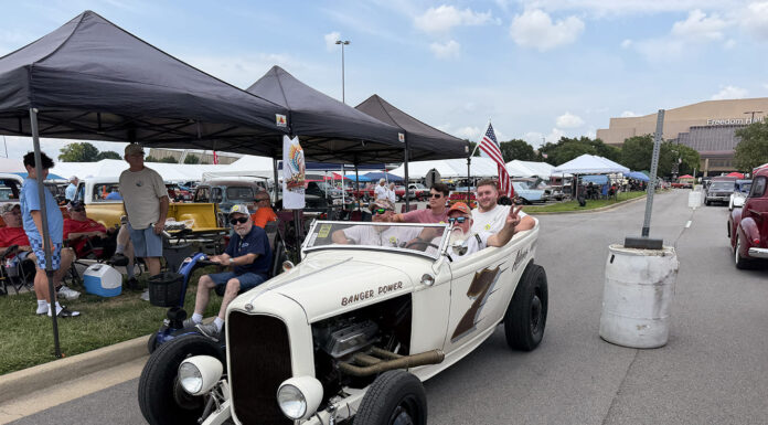 Front three-quarter view of an off-white 1932 Ford roadster hot rod with "Banger Power" hood lettering carrying passengers down the fairgrounds thoroughfare past spectator canopies.