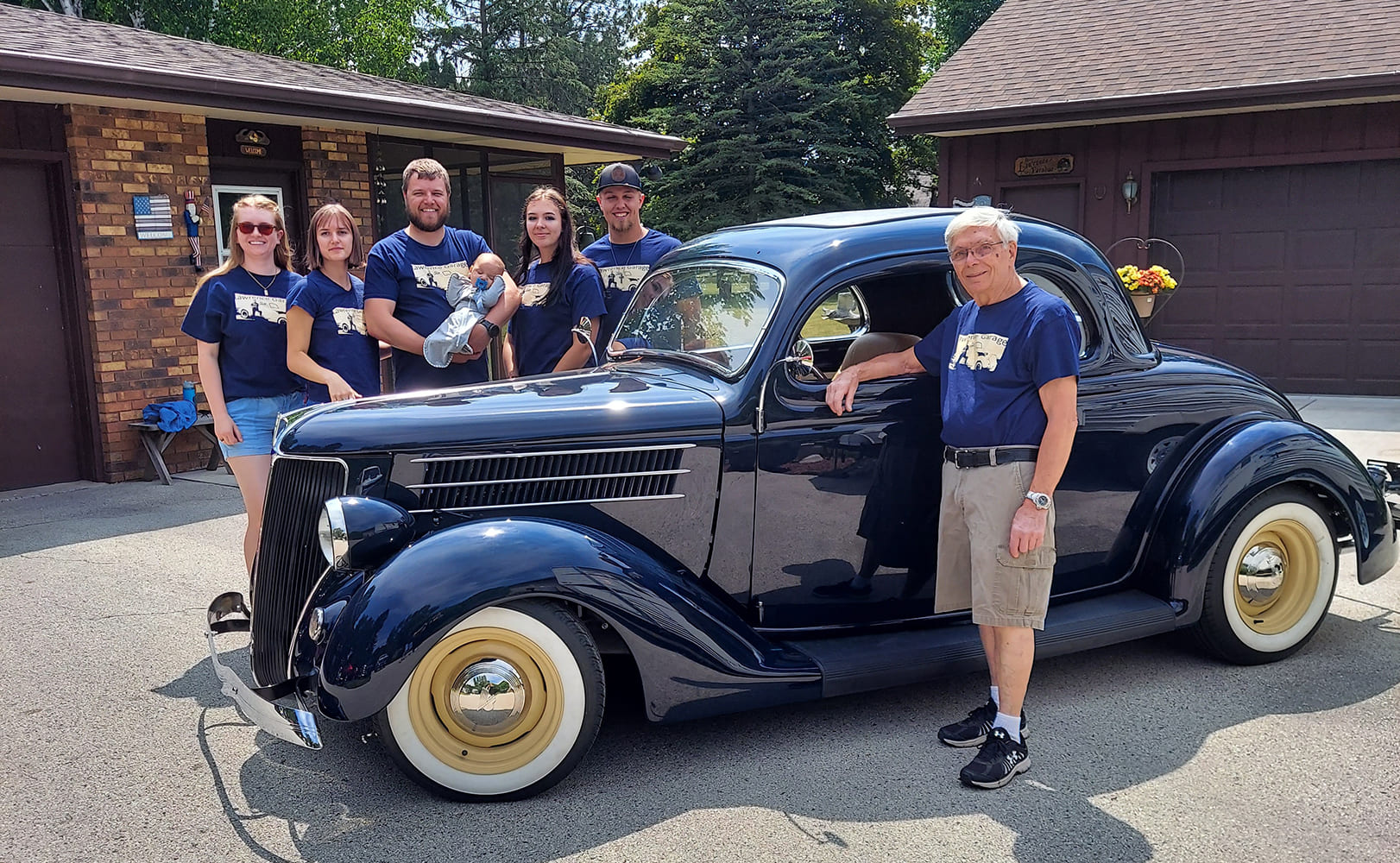 Tom Lawrence and his family, including grandchildren and a baby, posing proudly alongside his dark blue 1936 Ford five-window coupe in a paved driveway.