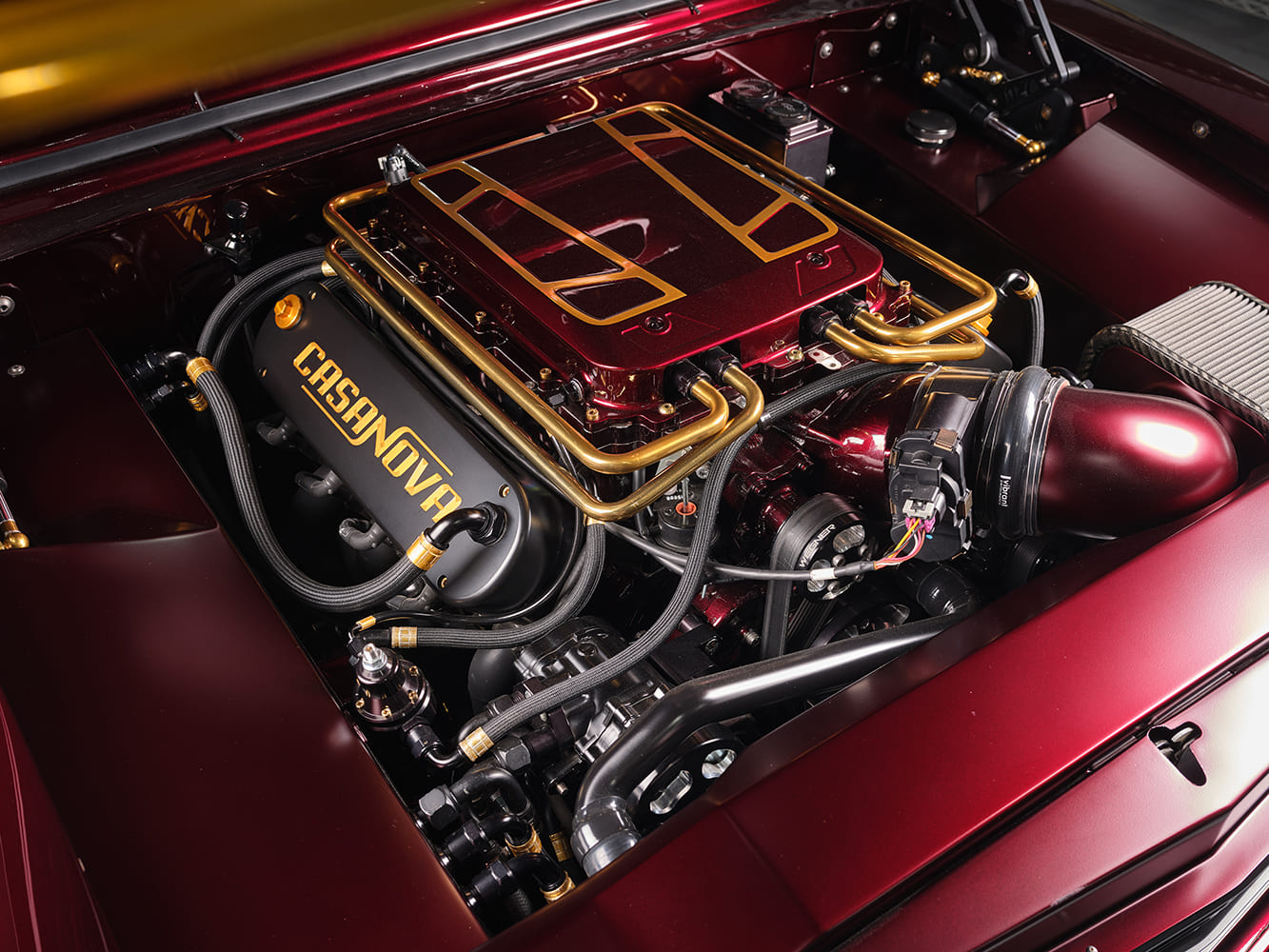 Overhead view of the clean custom engine bay in the 1967 Chevy Nova restomod with candy apple red intake, gold hard lines, Wegner front drive, and Casanova valve covers.