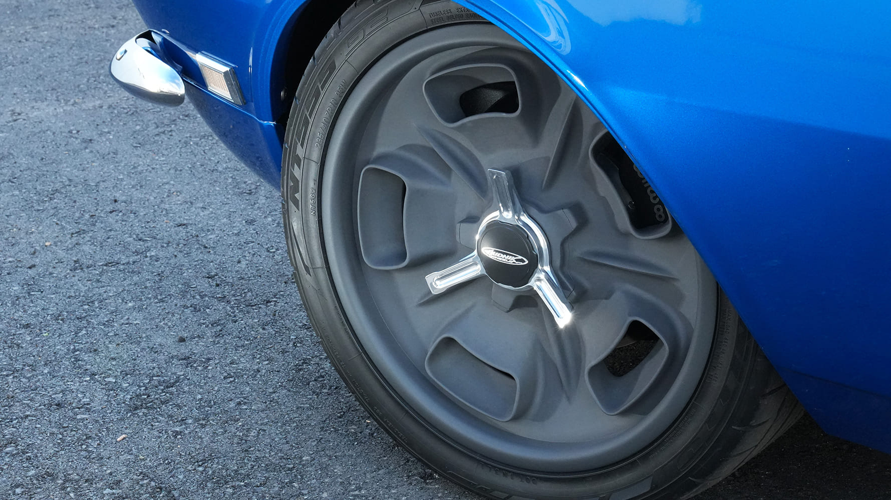 Close-up of a matte charcoal cast-style custom wheel with a three-ear polished knock-off spinner and low-profile performance tire on a blue 1968 Camaro.