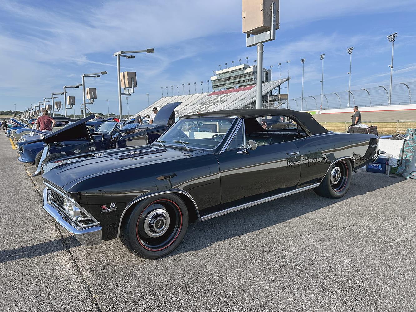 Wide outdoor shot of a glossy black 1966 Chevrolet Chevelle SS convertible with redline tires and dog-dish hubcaps parked on the infield of a racetrack.