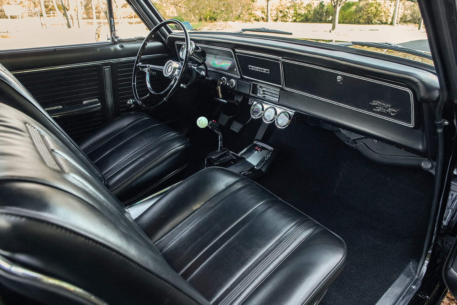 Driver-side interior view of a 1966 Chevy Nova SS featuring black vinyl bucket seats, column-mounted tachometer, auxiliary gauges, and manual floor shifter.