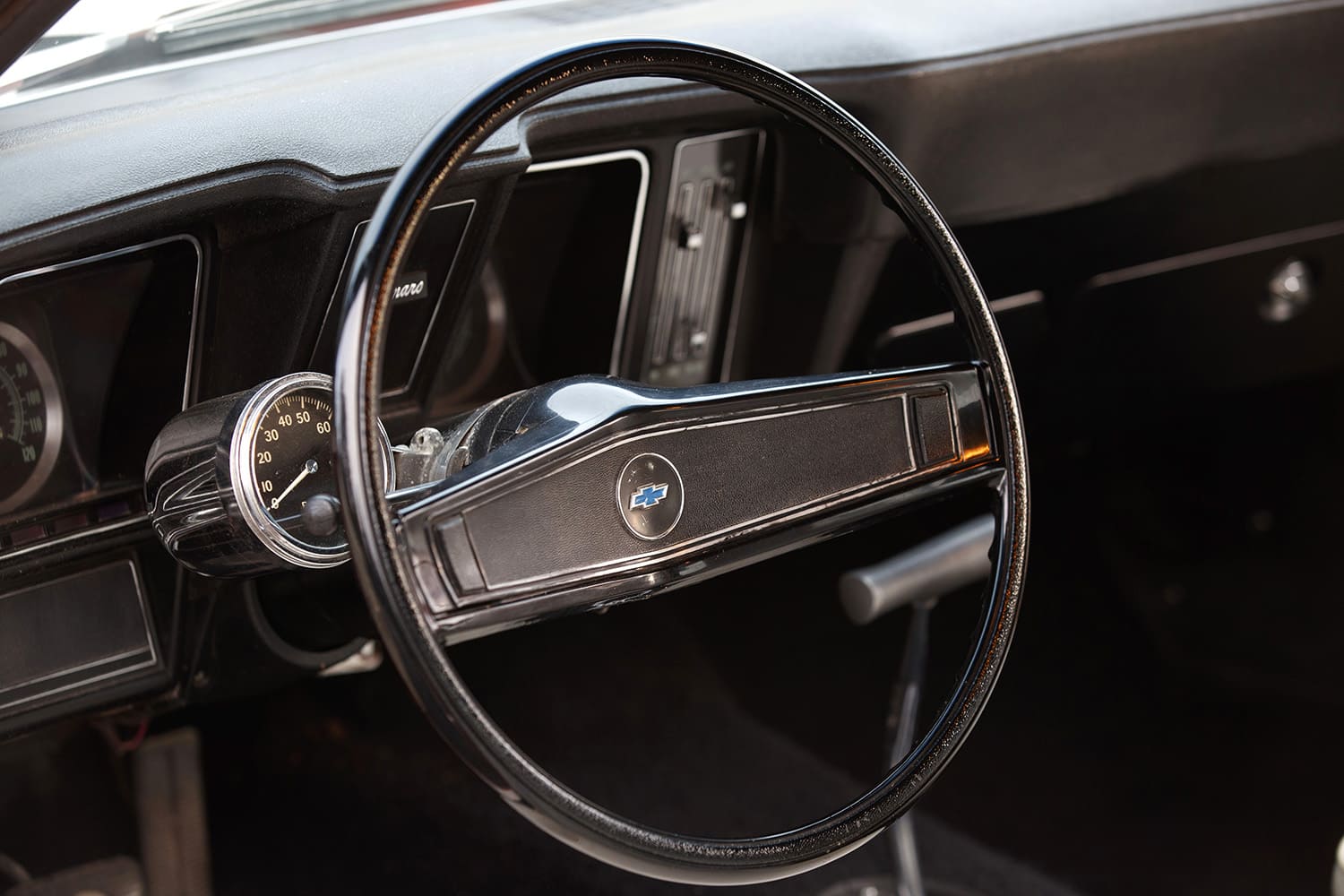 Driver perspective showing the factory two-spoke Chevrolet Bowtie steering wheel with a pedestal-mount Stewart-Warner tachometer attached to the steering column.