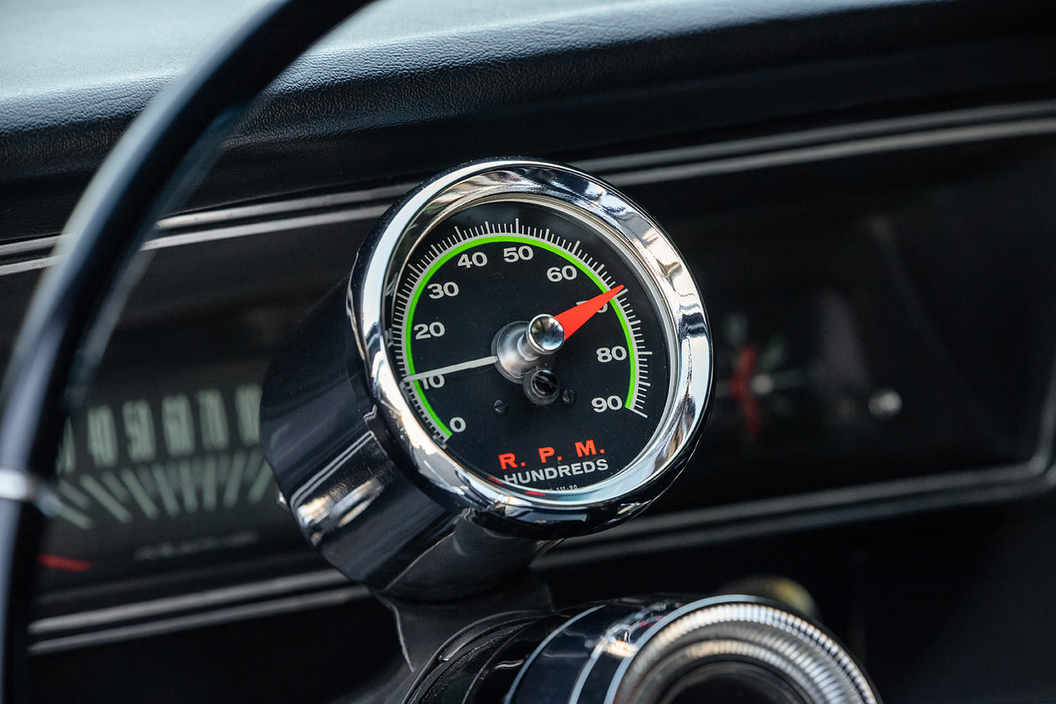 Close-up of a classic chrome-bezel Sun tachometer showing green accent markers and red needle mounted to the steering column.