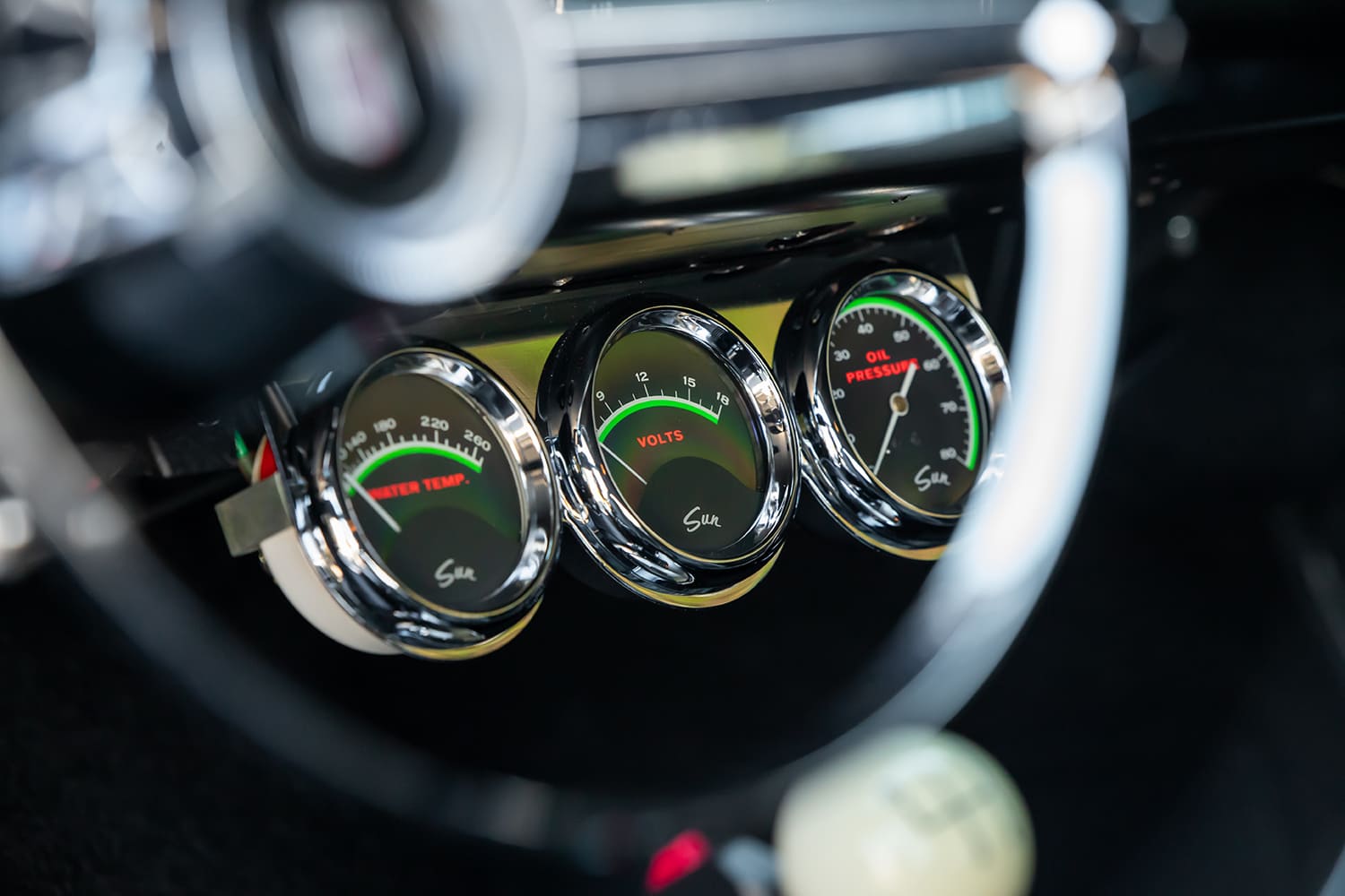 Macro view through the steering wheel of three chrome-housed Sun gauges monitoring water temperature, voltage, and oil pressure.