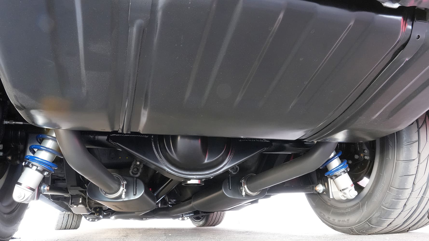 Underside view of the rear axle housing, dual exhaust system, fuel tank, and blue adjustable coilover shocks on a 1968 Camaro restomod.