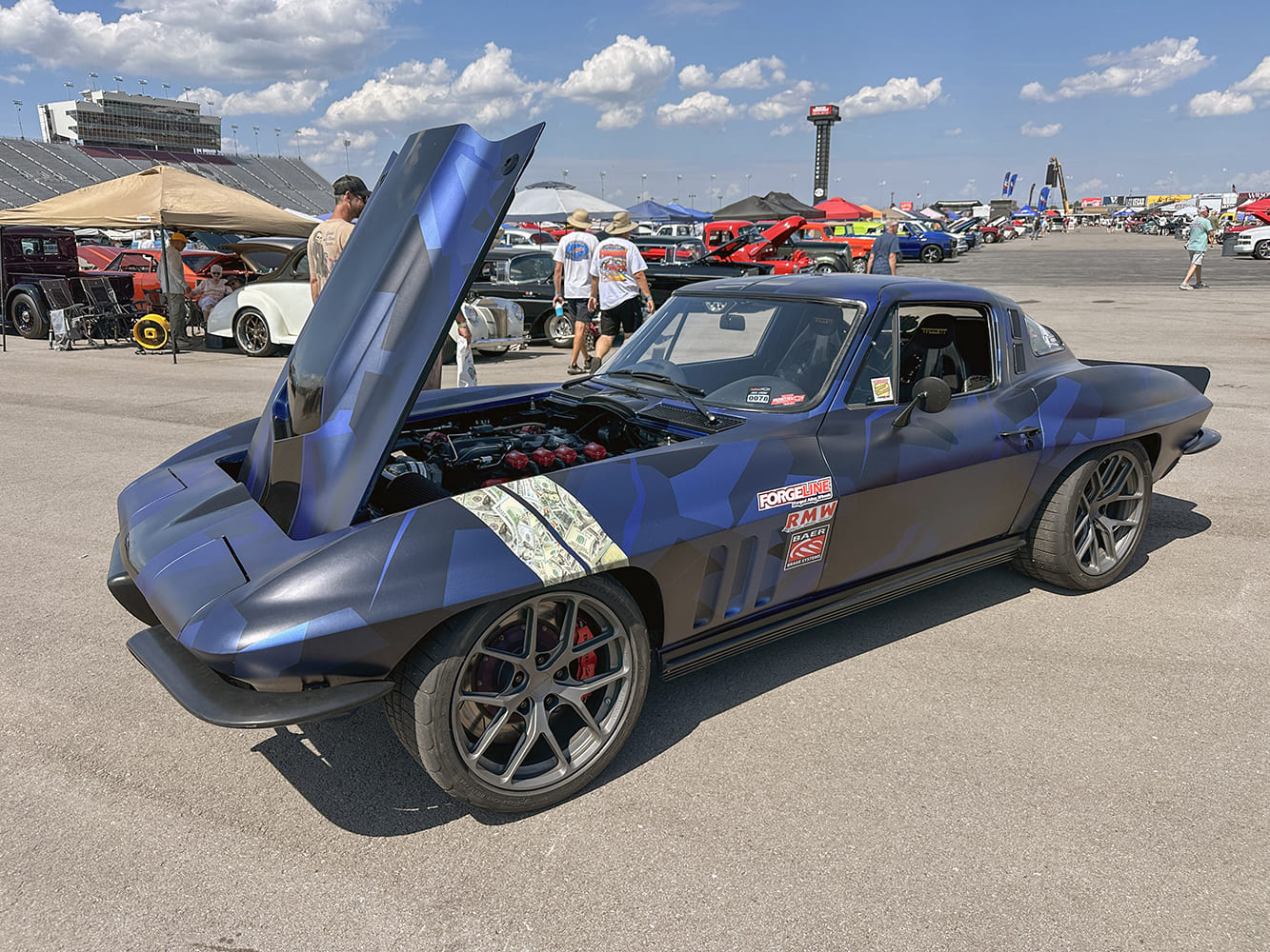 Front three-quarter view of a matte blue geometric camo Chevrolet Corvette C2 with modern multi-spoke wheels and open forward-tilting hood.