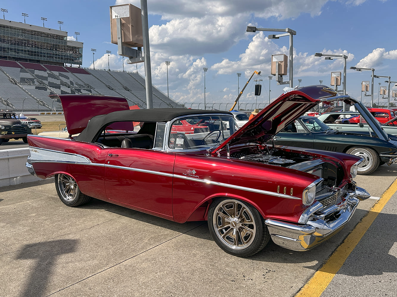 Metallic 57 Chevrolet Bel Air convertible with a black soft top, custom chrome wheels, and open hood and trunk on a speedway lot