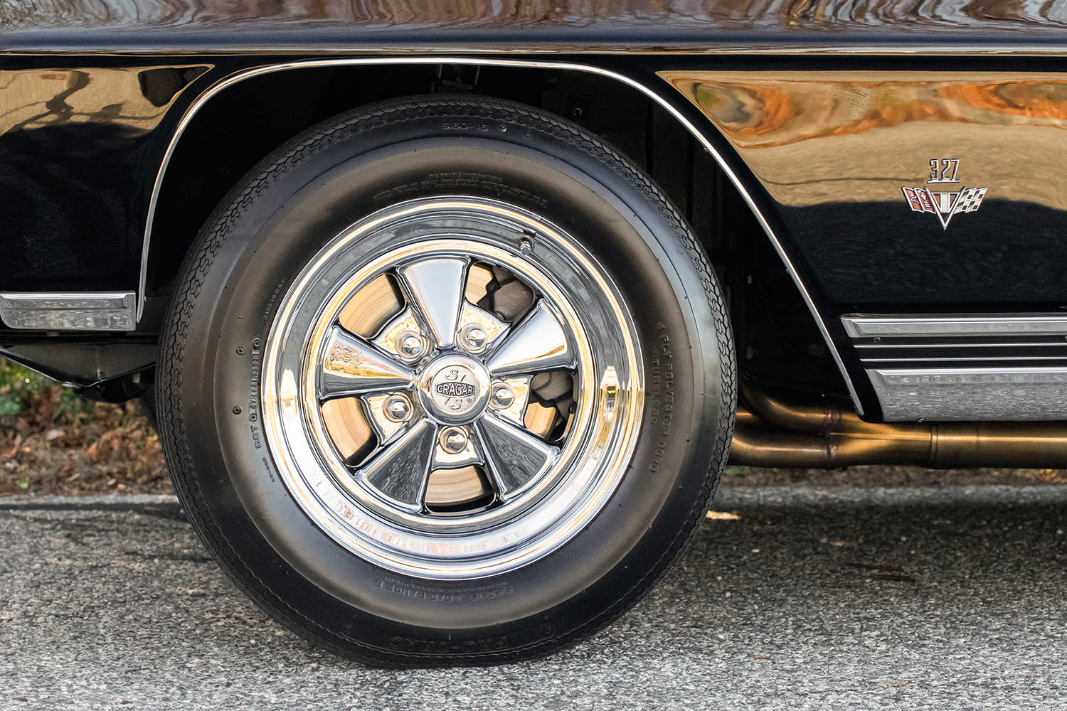 Side profile detail of a classic chrome Cragar S/S front wheel tucked under the front fender below an authentic Chevrolet 327 V8 cross-flag emblem.