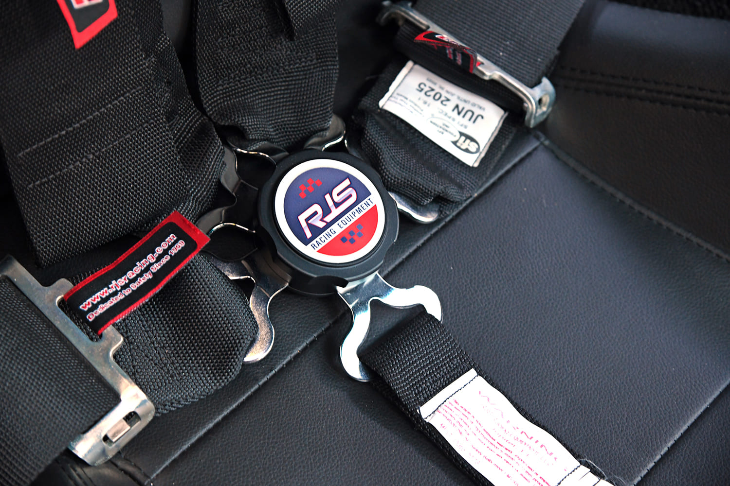 Close-up of an SFI-certified RJS Racing Equipment quick-release camlock harness buckle on black leather racing bucket seats.