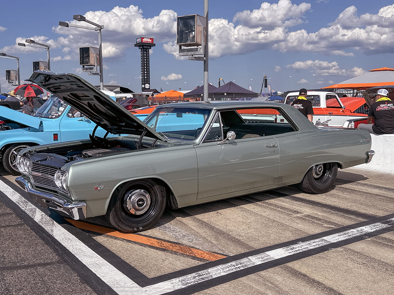 Side three-quarter angle of a muted gray-green 1965 Chevrolet Chevelle Malibu post car featuring an open hood and black steel wheels with chrome hubcaps.