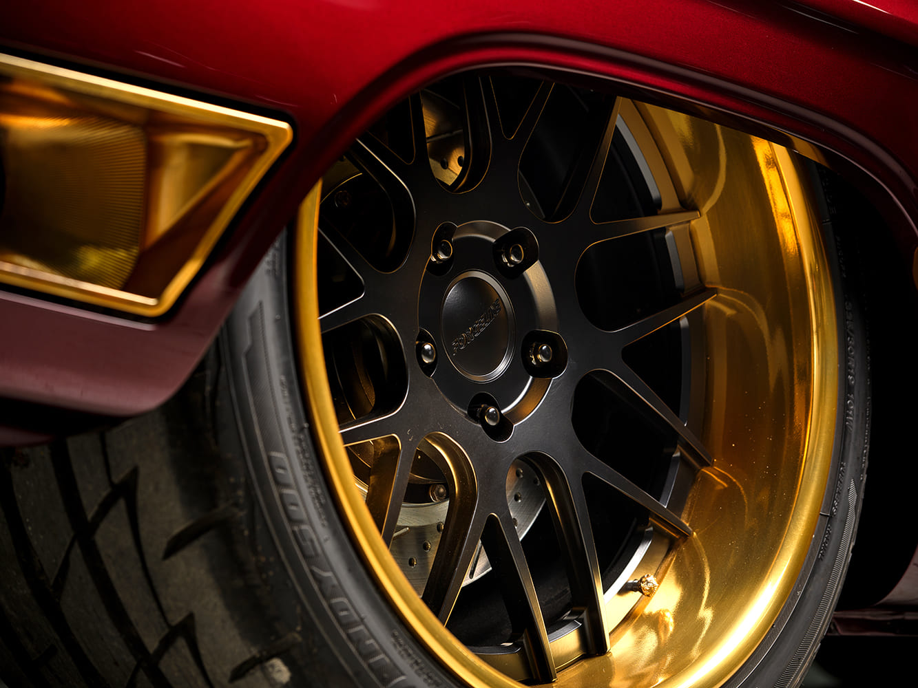 Close-up of a satin black Forgeline mesh wheel with a deep polished gold rim, low-profile performance tire, and drilled disc brake rotor.