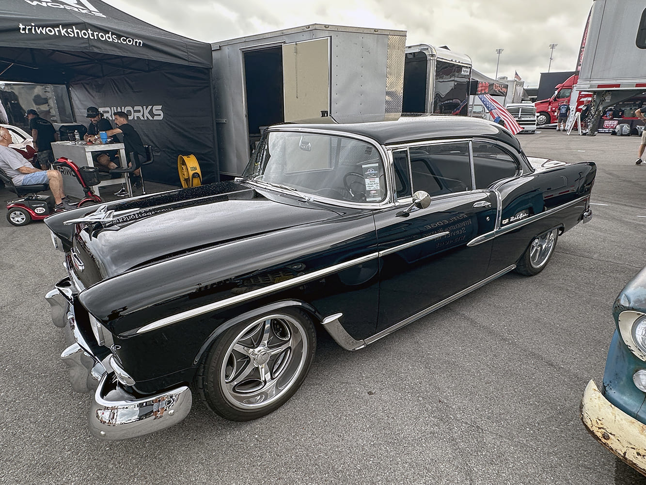 High front three-quarter view of a mirror-finish black 1955 Chevrolet Bel Air restomod with polished five-spoke wheels parked near event tents.