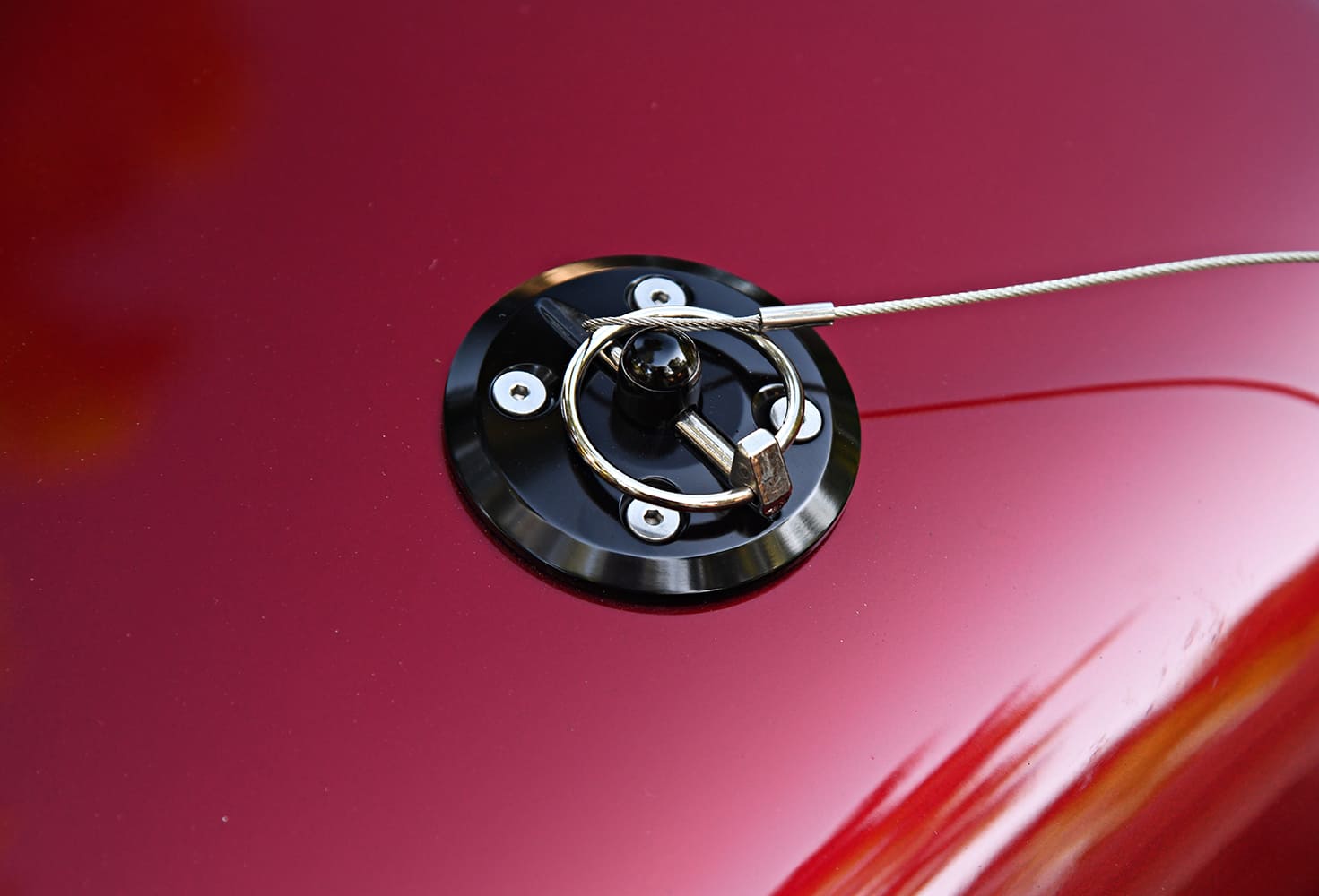 Extreme close-up of a quick-release lanyard hood pin and black anodized bezel installed on a metallic red fiberglass hood.