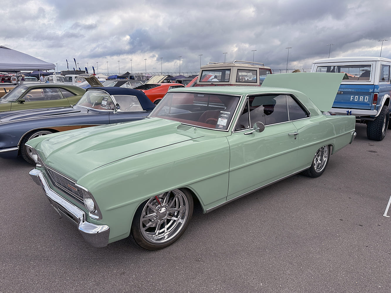 Low-angle front three-quarter shot of a mint green 1966 Chevy Nova hardtop featuring chrome multi-spoke wheels and an open trunk under an overcast sky