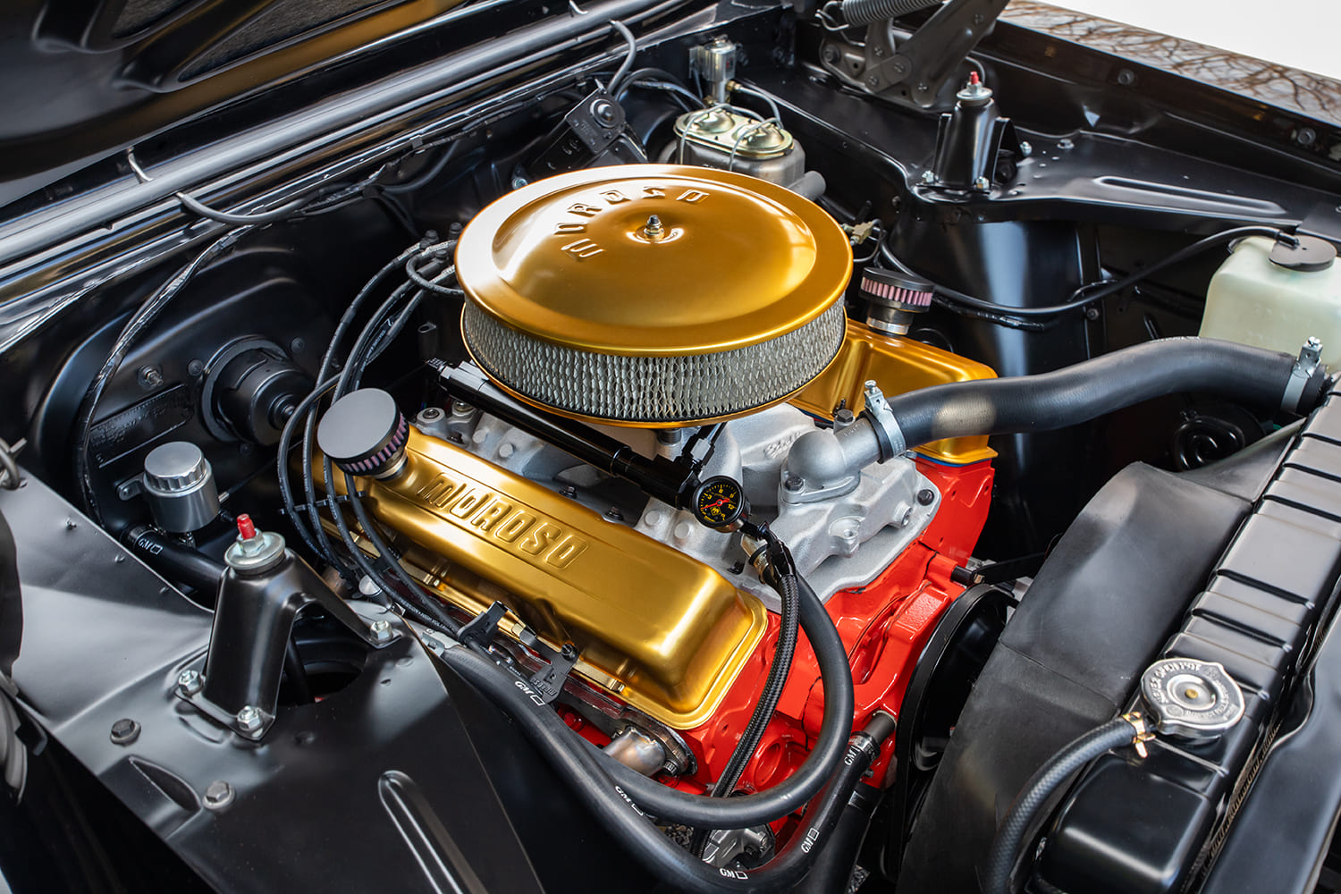 Angled view of a restored Chevrolet small-block V8 featuring an orange engine block, gold Moroso valve covers, matching gold air cleaner, and clean inner fenders.