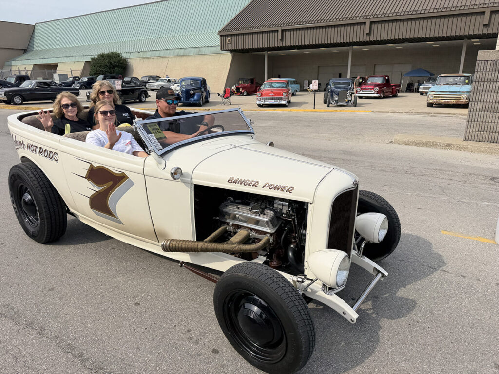 Elevated three-quarter view of a cream open-wheel roadster with race number 7 on the door carrying four smiling passengers across an asphalt lot at the street rod meet.