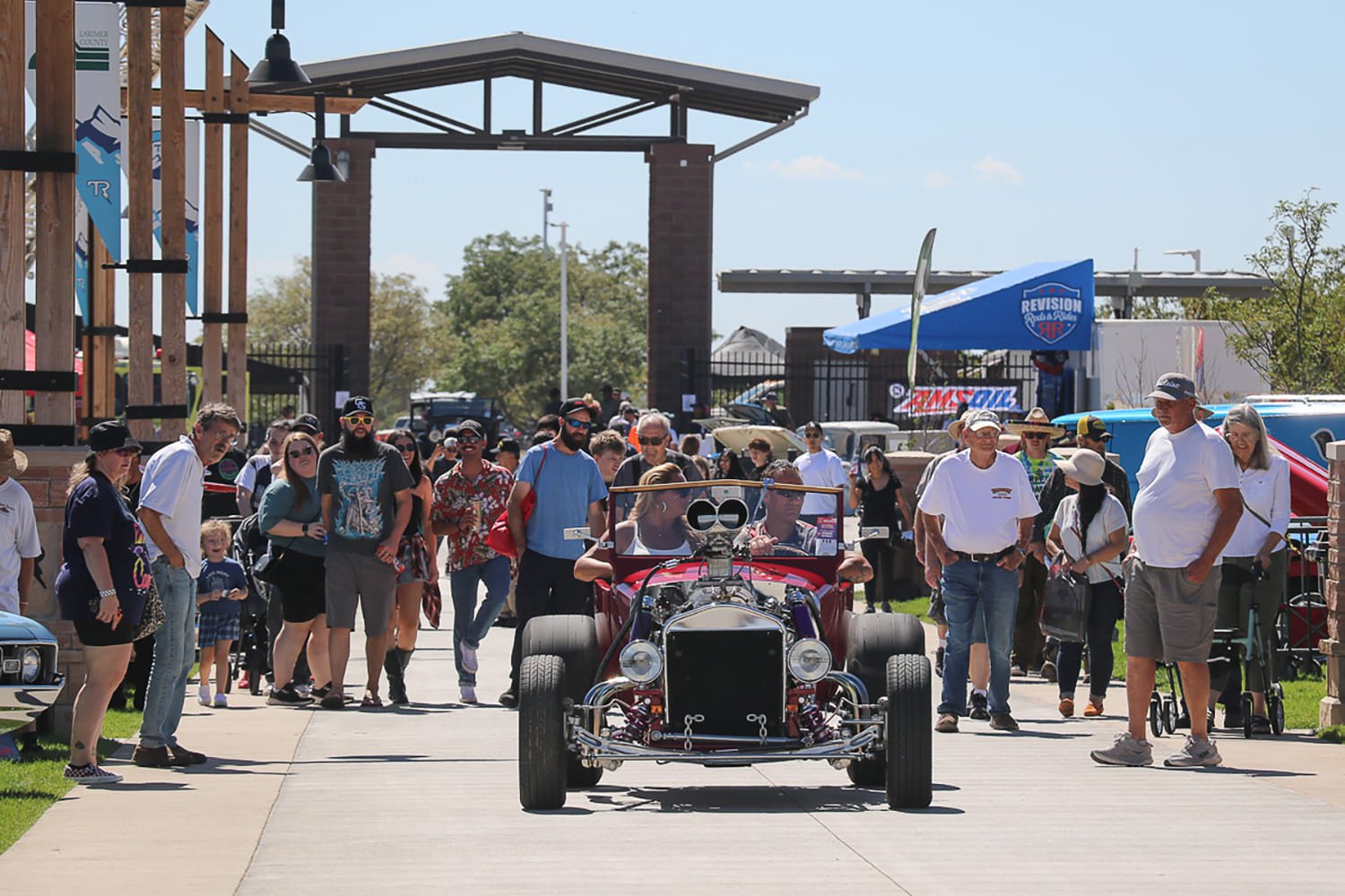 Red open-top T-bucket roadster hot rod with an exposed blower driving along a walkway crowded with spectators at the Good Guys Colorado Nationals car show in Loveland.