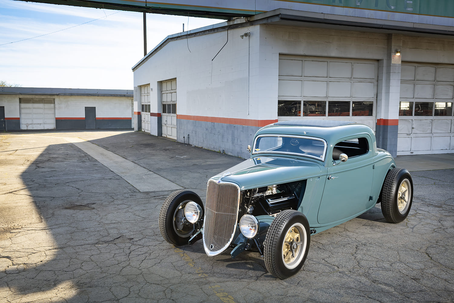 High front three-quarter view of a custom 1933 Ford Coupe hot rod with chrome vertical grille and open engine parked outside an automotive garage.