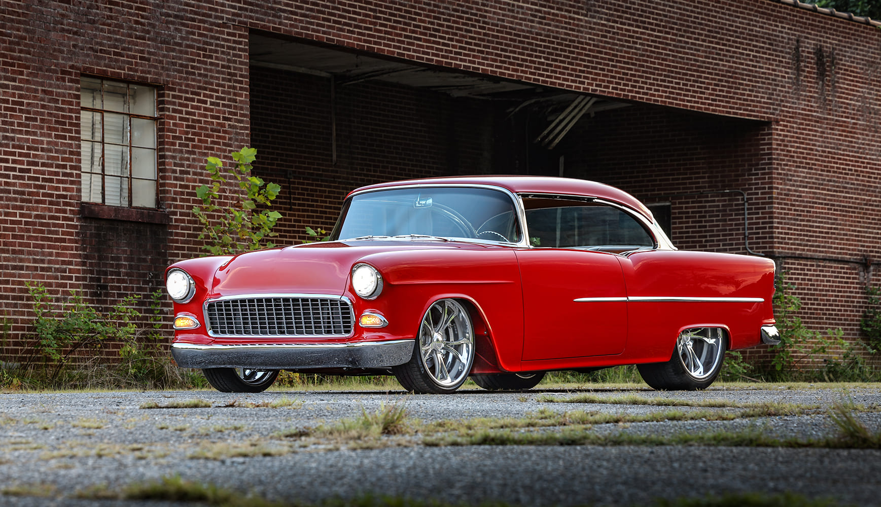 Front three-quarter angle of a shaved, custom red 1955 Chevrolet Bel Air hardtop parked in front of an old brick building.