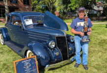 Car owner Tom Lawrence holding a tall multi-tier car show trophy next to his dark blue 1936 Ford coupe displayed with its hood open on grass.