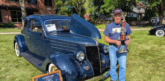 Car owner Tom Lawrence holding a tall multi-tier car show trophy next to his dark blue 1936 Ford coupe displayed with its hood open on grass.