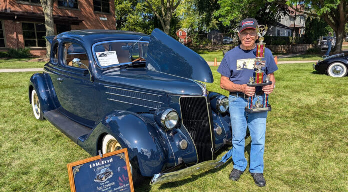 Car owner Tom Lawrence holding a tall multi-tier car show trophy next to his dark blue 1936 Ford coupe displayed with its hood open on grass.