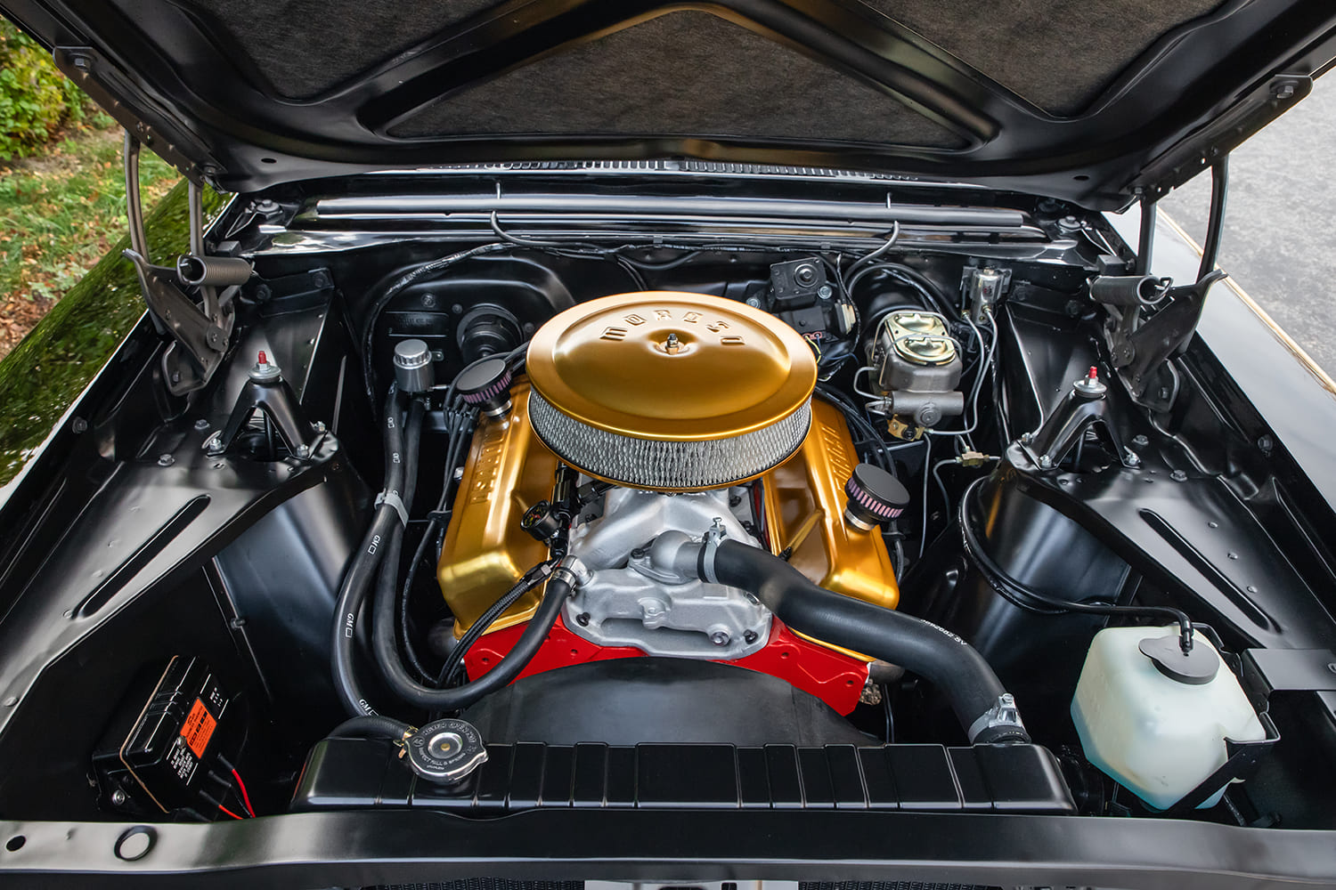 Wide top-down view of the entire gloss black engine bay showcasing a gold-accented small-block V8, aluminum intake, and brass-capped dual master cylinder.
