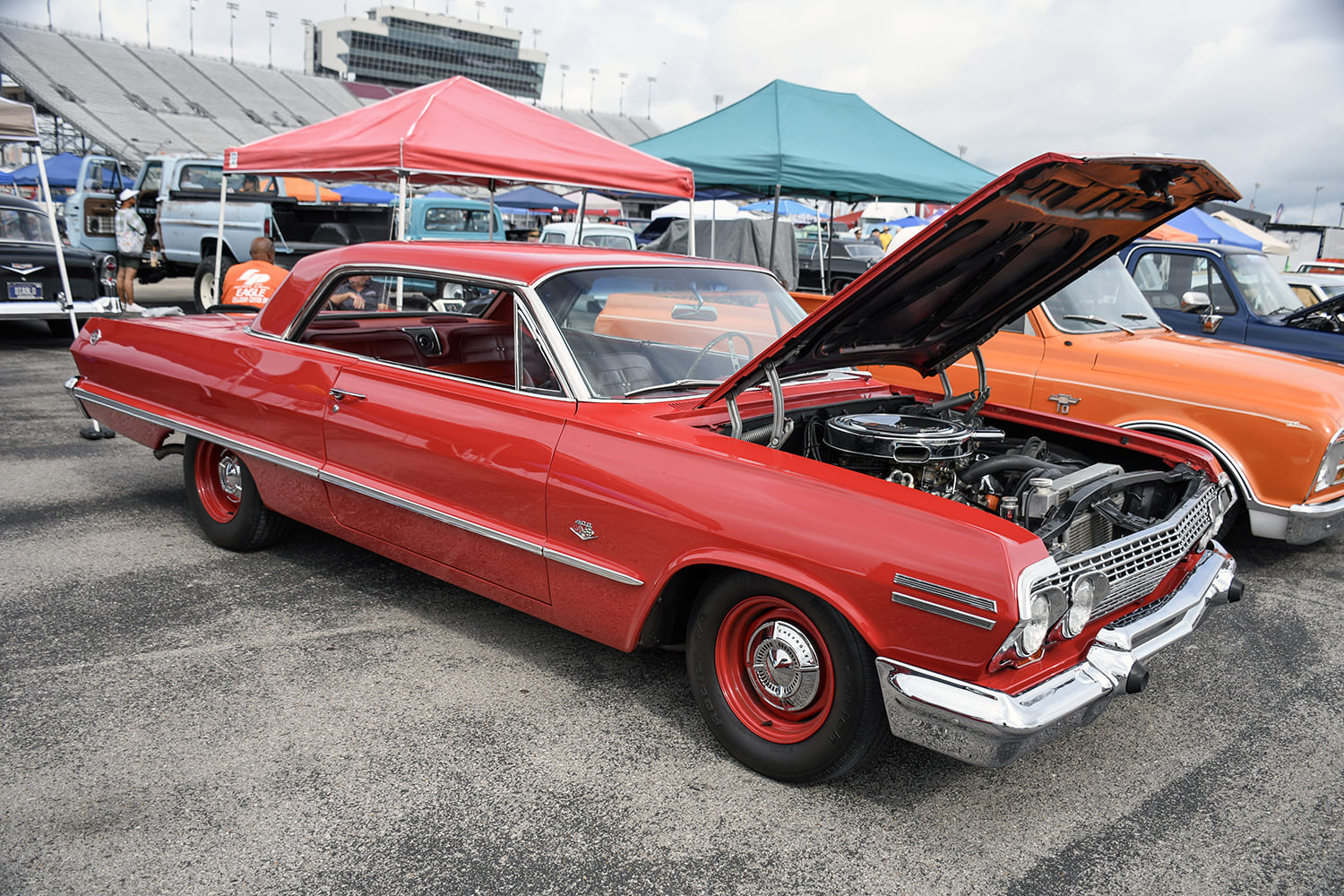 Front three-quarter view of a bright red 1963 Chevrolet Impala hardtop with an open hood, red steel wheels with dog-dish hubcaps, and full chrome trim at an outdoor speedway car show.
