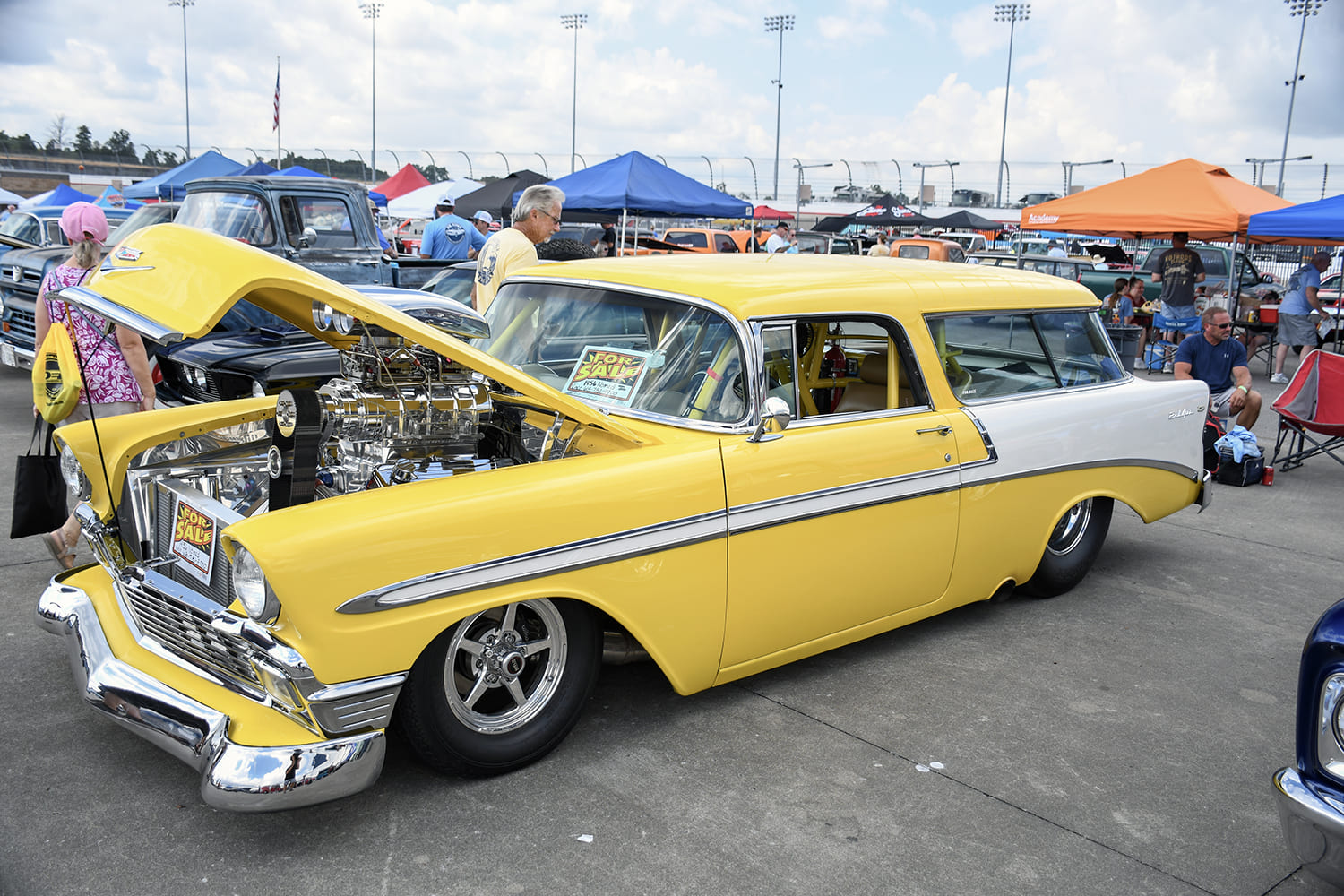 right yellow and white 1956 Chevrolet Nomad wagon showing a massive polished supercharger rising out of the engine bay, parked on speedway asphalt.