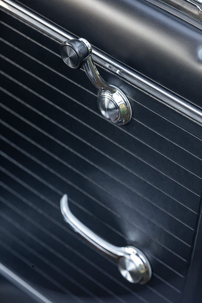Close-up of the chrome window crank, black textured vinyl door panel, and chrome interior door release handle on a 1966 Chevy Nova.