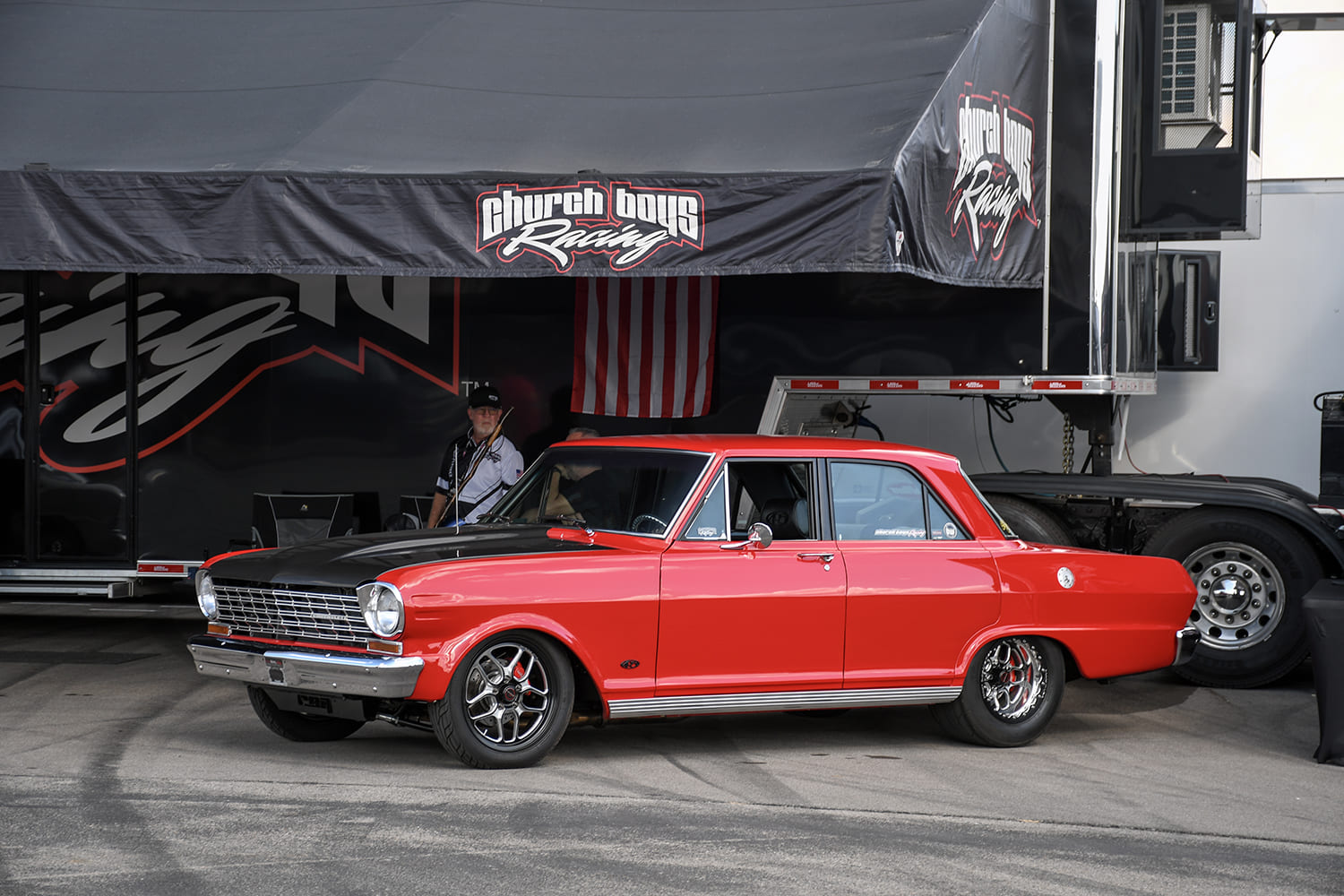 Side view of a red 1964 Chevrolet Chevy II Nova four-door sedan with a black hood and lightweight racing wheels parked in front of a Church Boys Racing hauler awning.