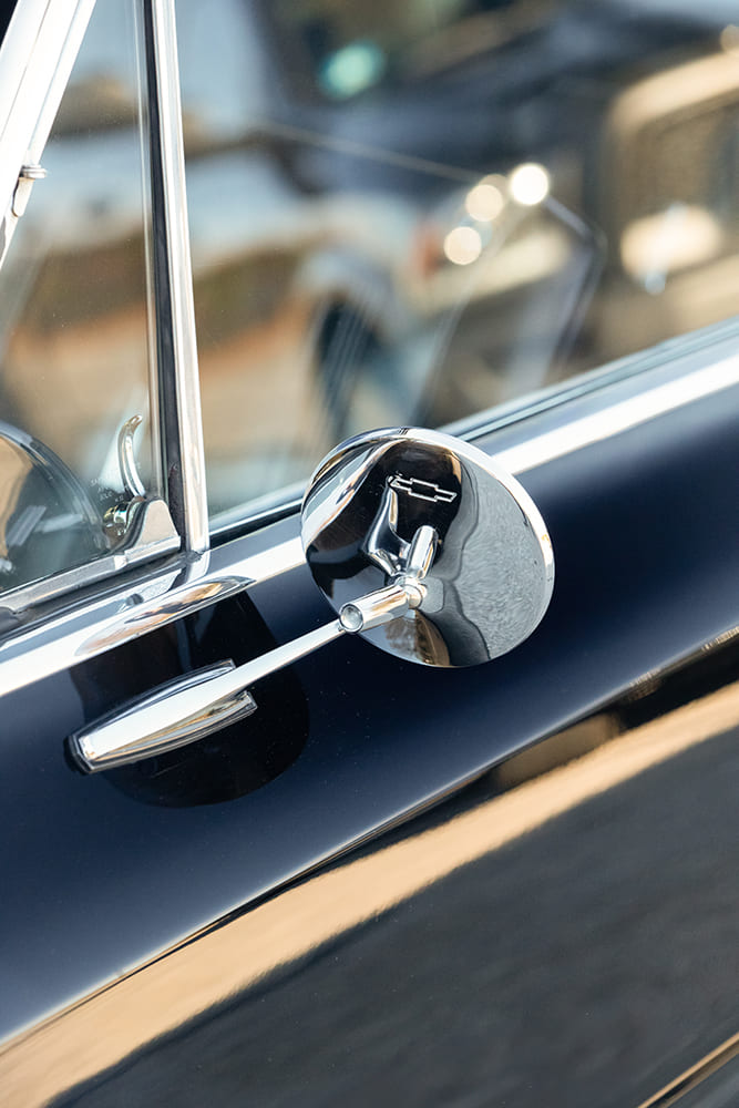 Exterior detail of a round chrome side mirror embossed with the Chevrolet Bowtie logo on the door of a black 1966 Chevy Nova.