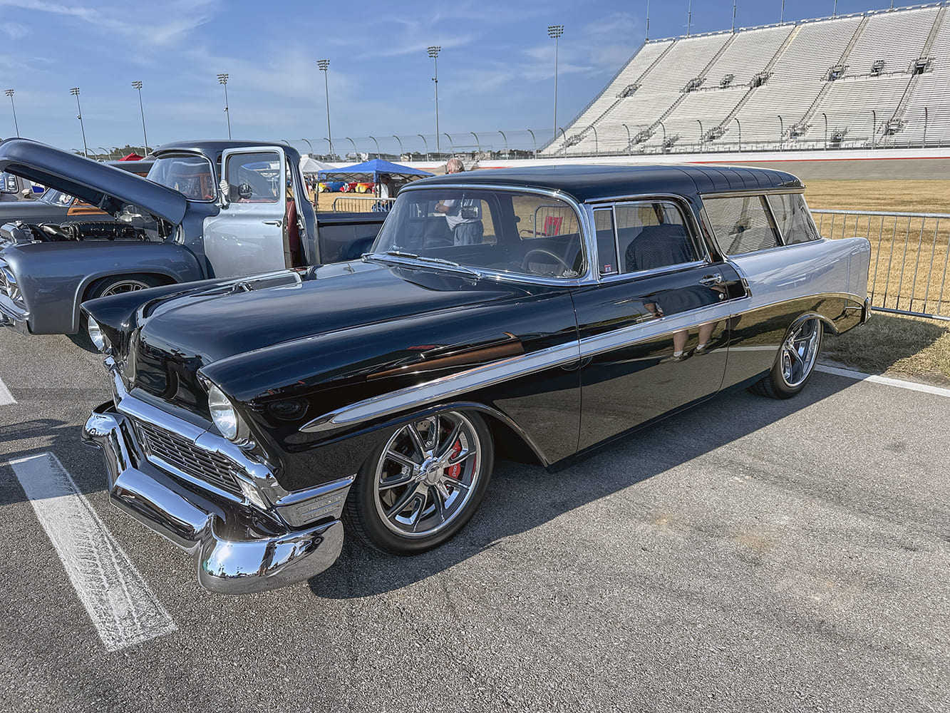 Front three-quarter view of a customized black and silver two-tone 1956 Chevrolet Nomad wagon riding low on modern polished chrome wheels in front of grandstand seating.