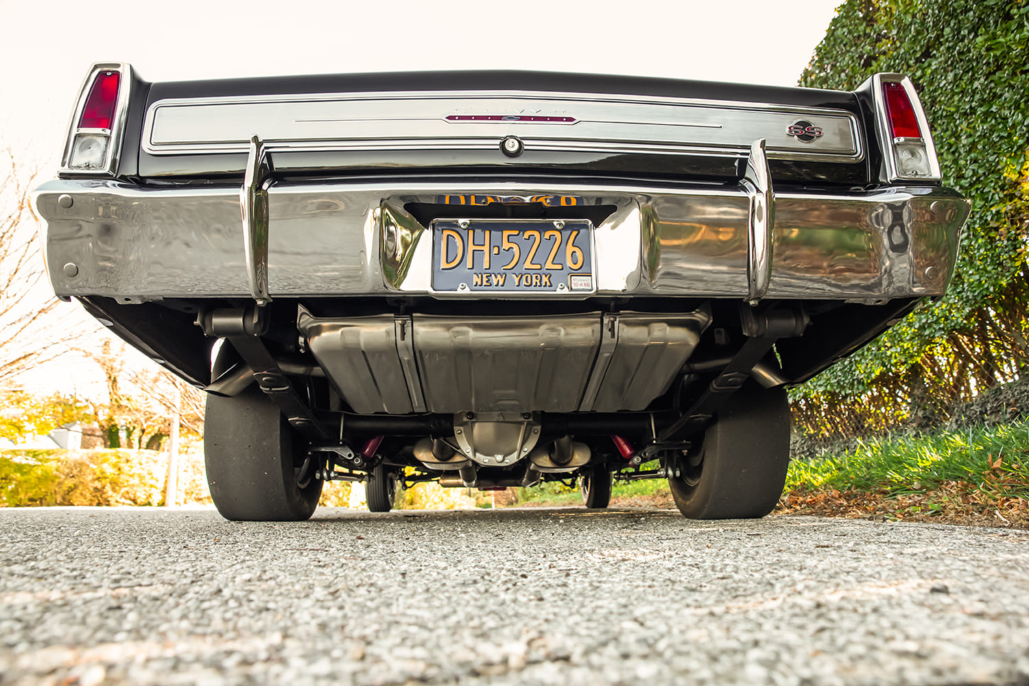Low ground-level view under the rear bumper of a 1966 Chevy Nova showing the fresh fuel tank, rear differential, traction bars, and wide rear tires.
