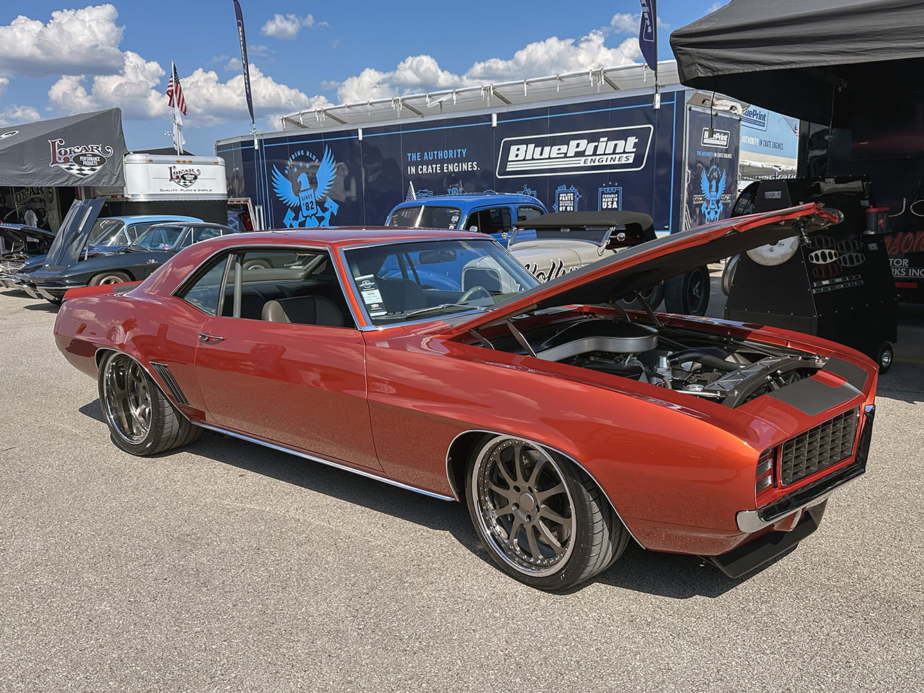 Front three-quarter perspective of a metallic burnt copper 1969 Chevrolet Camaro coupe with an open cowl hood and charcoal gray wheels parked outside vendor displays.