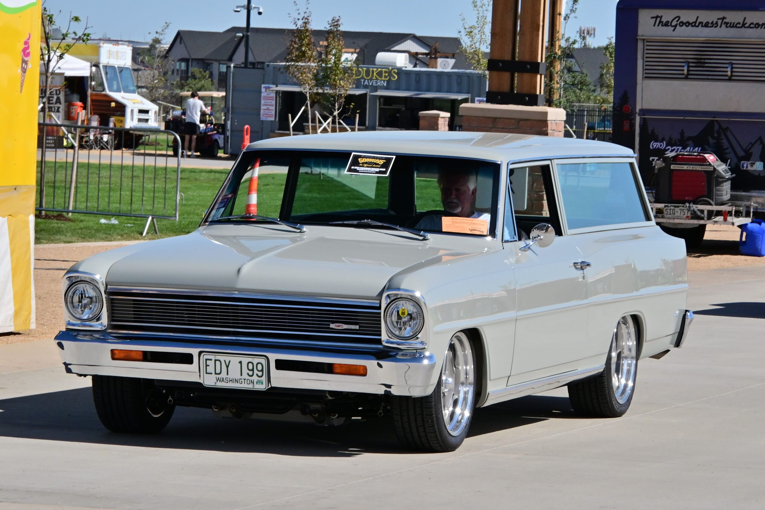 Front three-quarter angle of a sleek light-gray two-door Chevy II Nova station wagon driving slowly across the show grounds under bright sunlight.