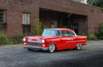 Slammed 1955 Chevy Bel Air coupe in gloss red paint parked on pavement outside an industrial brick warehouse.