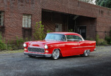 Slammed 1955 Chevy Bel Air coupe in gloss red paint parked on pavement outside an industrial brick warehouse.