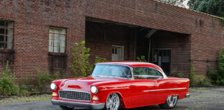 Slammed 1955 Chevy Bel Air coupe in gloss red paint parked on pavement outside an industrial brick warehouse.