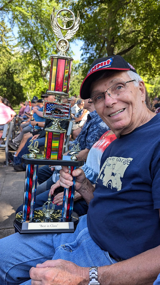 Close-up of smiling car owner in a Summit Racing cap holding his "Best in Class" trophy while seated outdoors at a car show award ceremony.