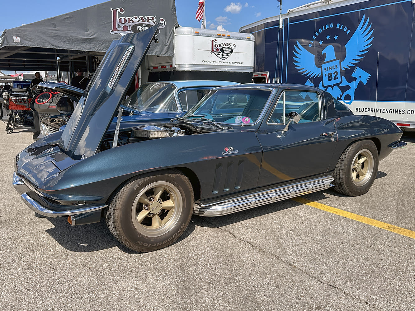 Side three-quarter profile of a deep charcoal blue 1966 Chevrolet Corvette C2 Sting Ray coupe featuring side exhaust pipes, gold vintage wheels, and an open front-tilt hood.