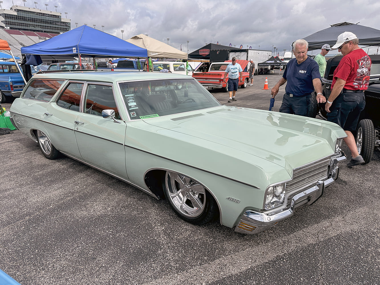 Front three-quarter high angle of a lowered pale sage green 1970 Chevrolet Kingswood 400 wagon on chrome billet wheels with spectators gathered nearby.