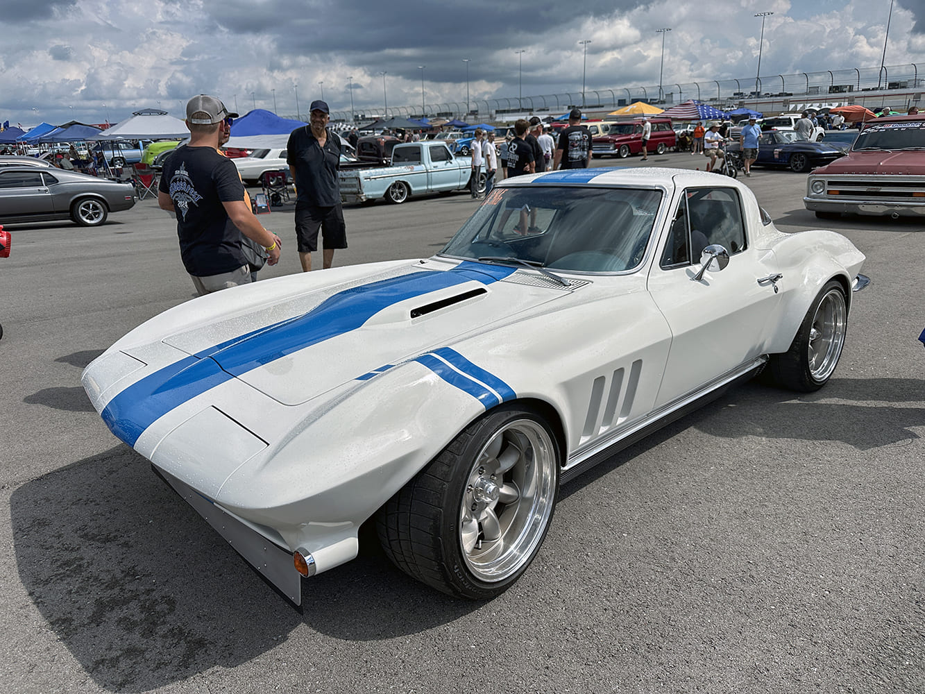 High front three-quarter view of a widebody white C2 Corvette coupe with dual blue racing stripes, deep dish wheels, and front air splitter at a car show.