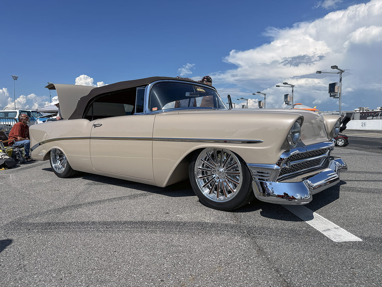 Ground-level view of a slammed champagne beige 1956 Chevrolet Bel Air convertible with a brown cloth top and full chrome wire-style wheels.