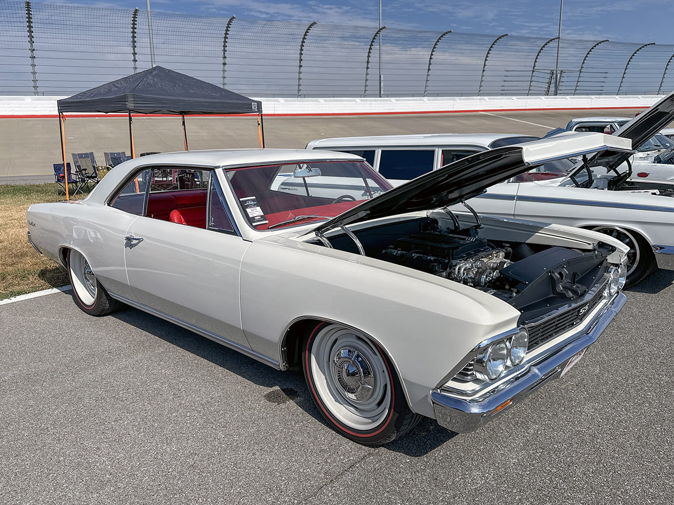 High front three-quarter perspective of a pearl white 1966 Chevelle SS showing a red leather interior, modern engine under the open hood, and redline steel wheels.