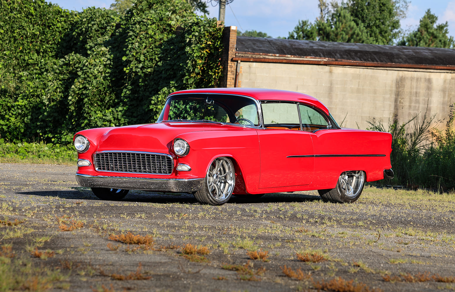 Driver-side front three-quarter view of a custom red 1955 Bel Air with billet wheels and shaved badges against green foliage.
