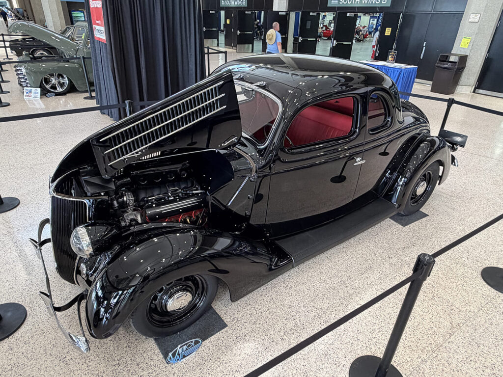 High front three-quarter view of a gloss black 1936 Ford street rod coupe featuring an open louvered hood, modern fuel-injected engine, and deep red interior.